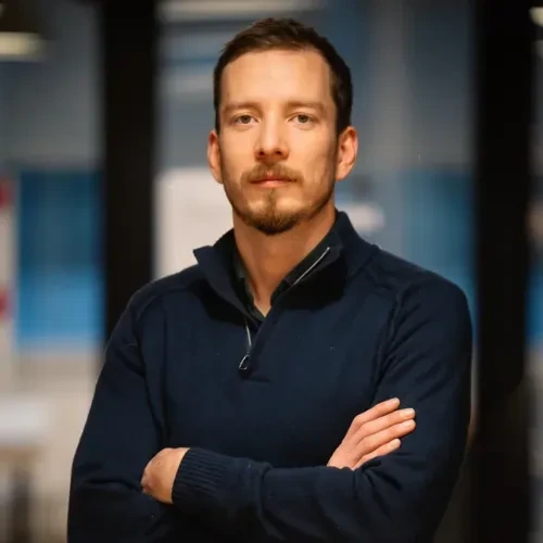 Portrait of Vitaly Pecherskiy with arms crossed in a dark sweater, standing indoors against a blurred background.